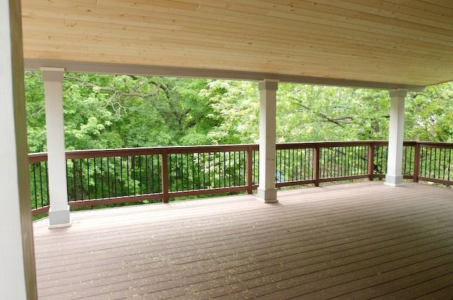 Covered wooden deck with white columns and brown railing, overlooking a green wooded area.
