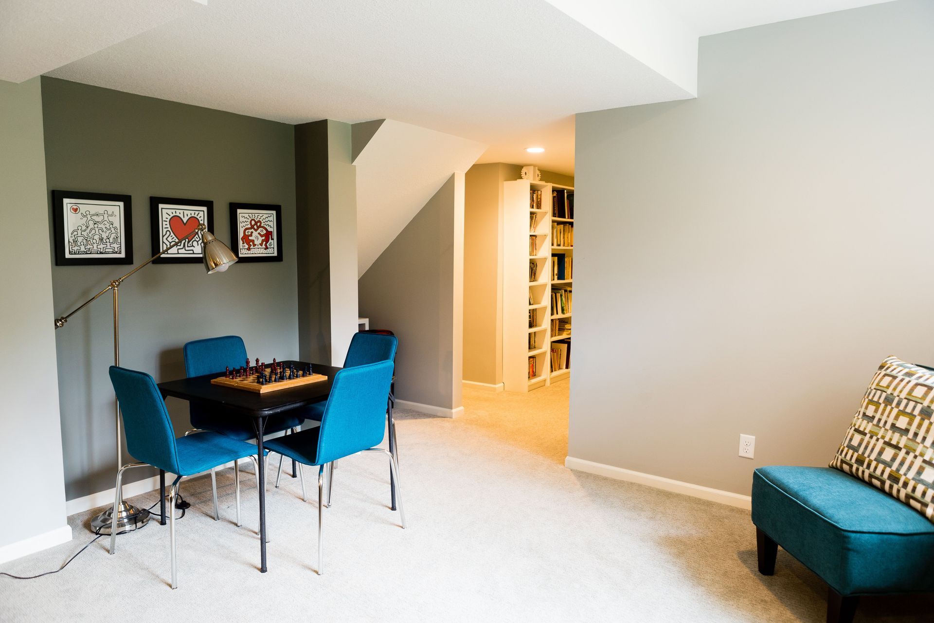 A cozy basement with a chess table, blue chairs, art, and a bookcase.