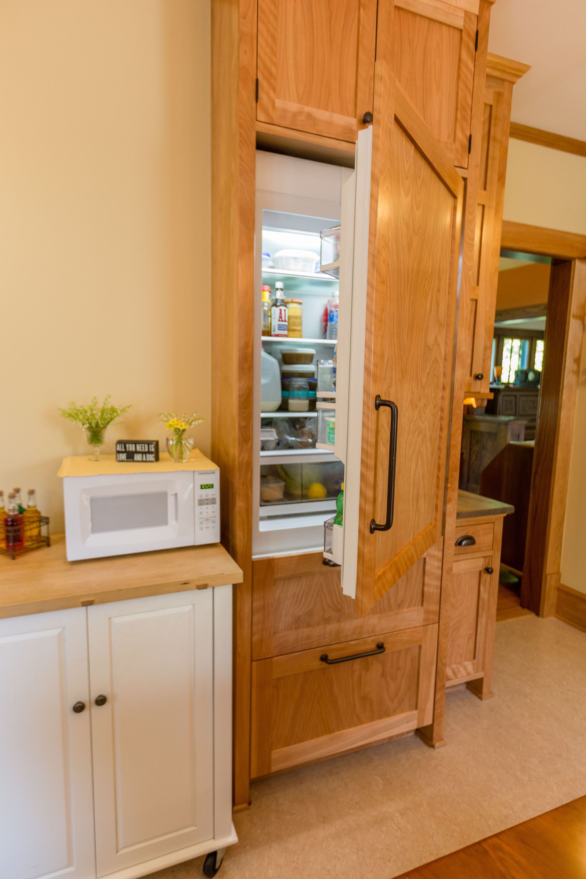 Open, wooden-paneled refrigerator with food inside. A microwave sits on a white cabinet.