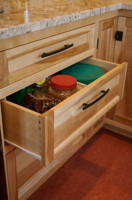 Open kitchen drawer, light wood, holding food containers and bags. Black handle, granite countertop, red floor.