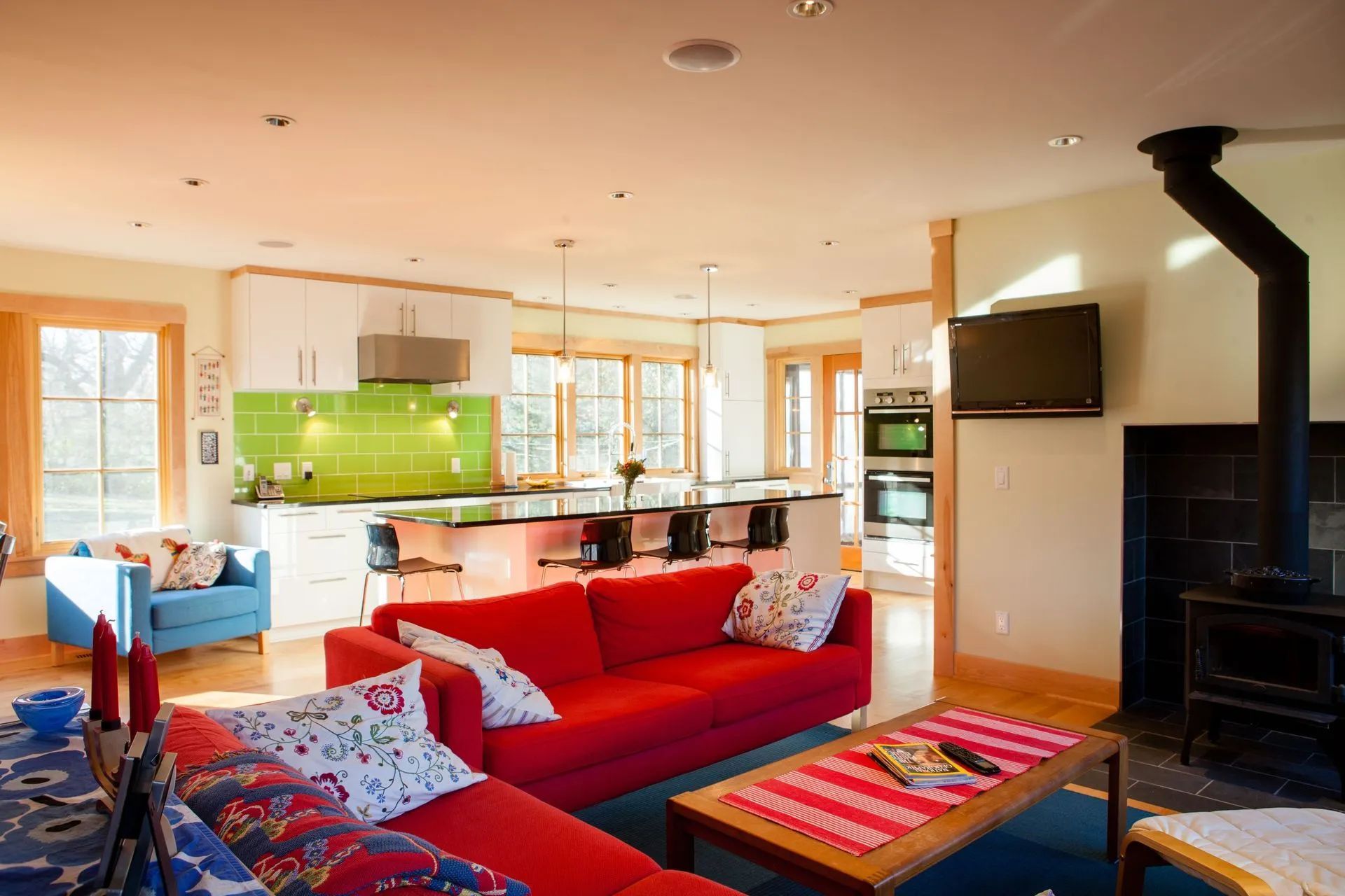 Bright open-plan living space with red sofa, blue armchair, and kitchen with green backsplash.