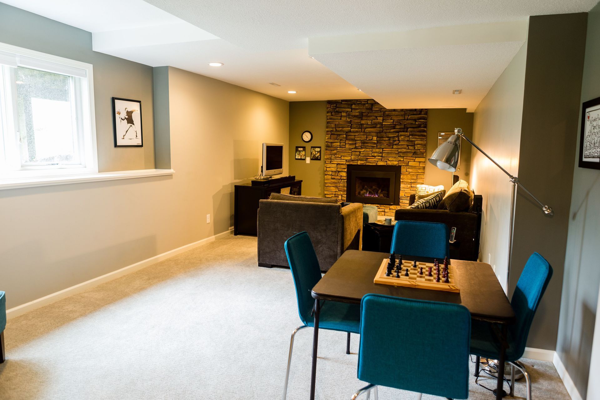 Basement living room with fireplace, game table, and teal chairs. Neutral walls, stone fireplace surround.