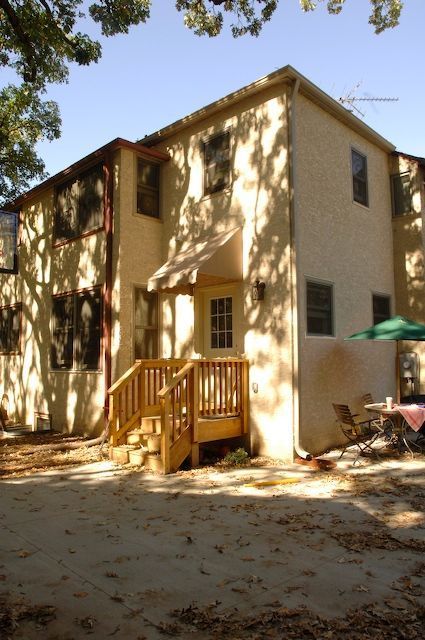 Two-story stucco building with wooden stairs and a door with an awning; shadows cast by trees.