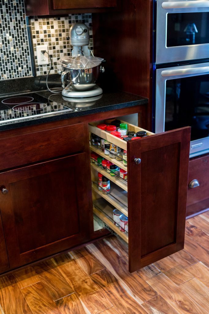Dark wood kitchen cabinet with a pull-out spice rack filled with jars. A double oven is visible.
