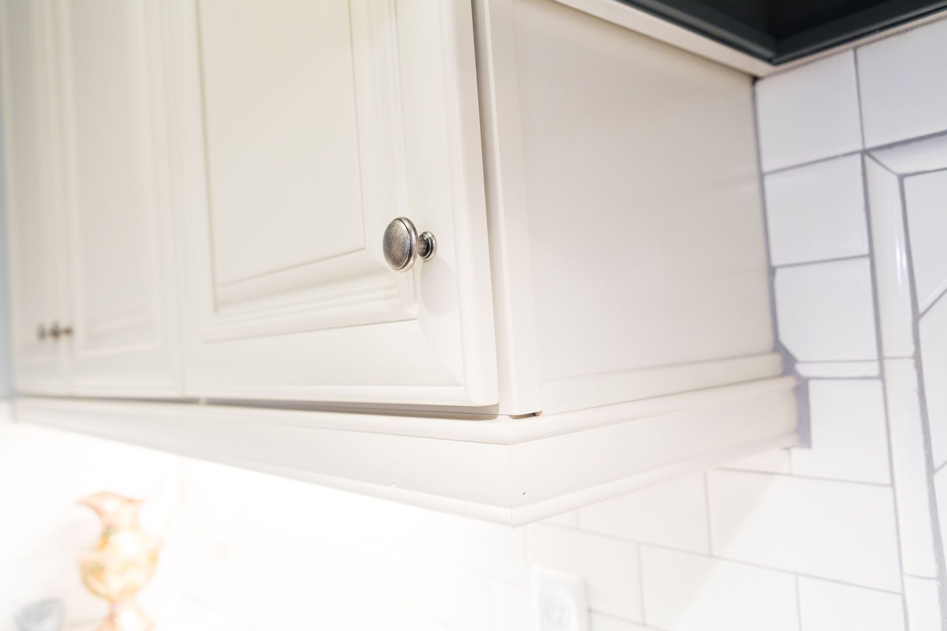 White kitchen cabinet with silver knob, mounted above white tile backsplash.