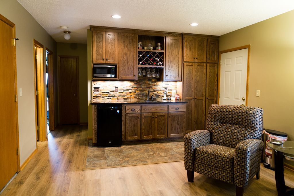 A home bar with wooden cabinets, a microwave, and a small refrigerator. A patterned armchair sits nearby.