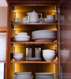 Wooden cabinet with illuminated shelves holding white dishware, including plates, bowls, cups, and a pitcher.