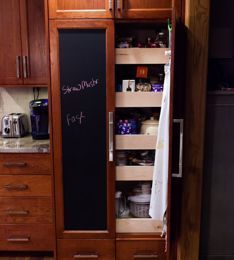 Kitchen pantry with pull-out shelves and chalkboard door. Contains food items and a hanging towel. Brown wood.