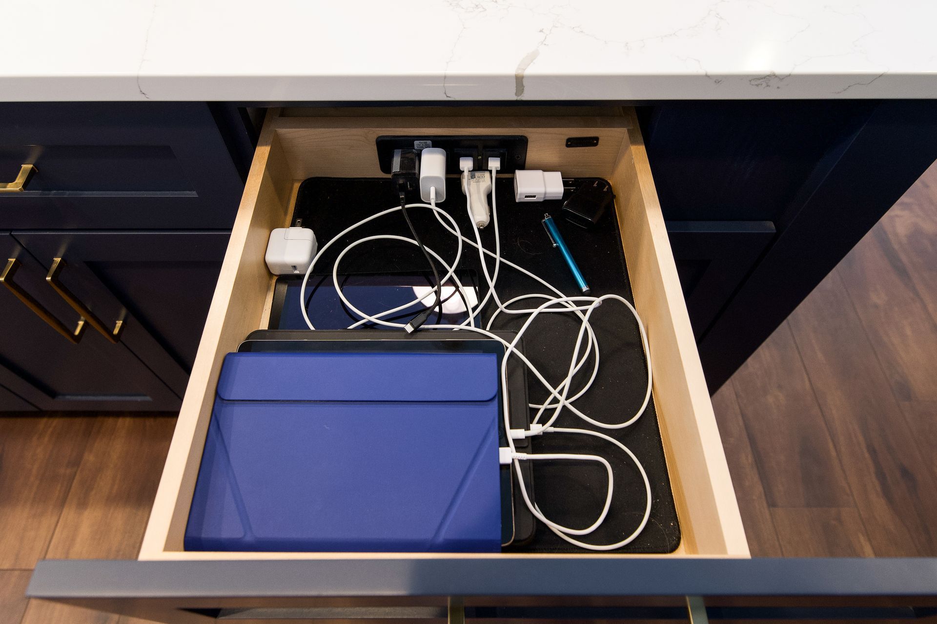 Drawer with chargers, cables, and a blue tablet case inside a kitchen cabinet.