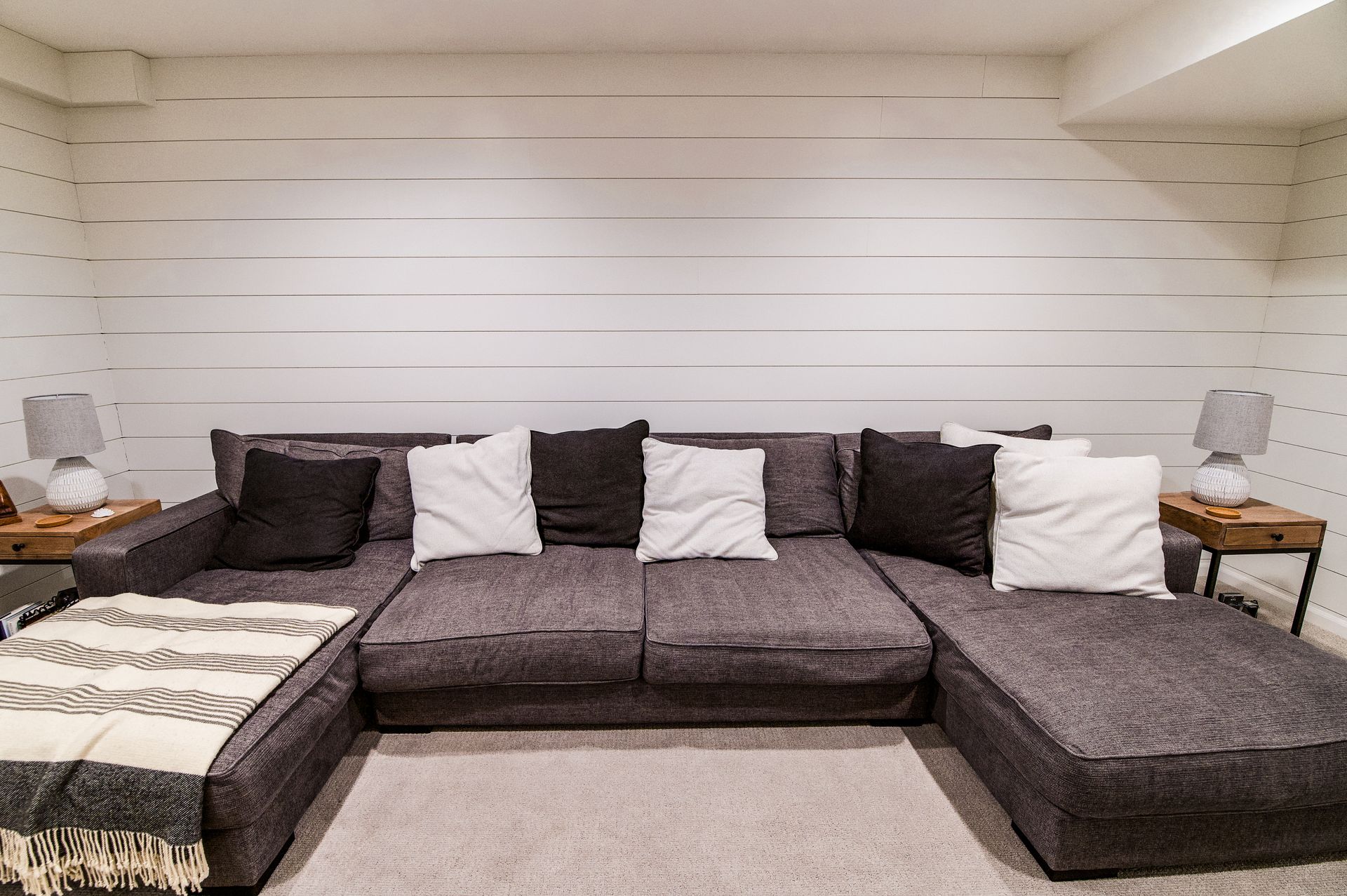 Gray sectional sofa with white and black pillows, wood side tables, and lamps against a horizontal plank wall.