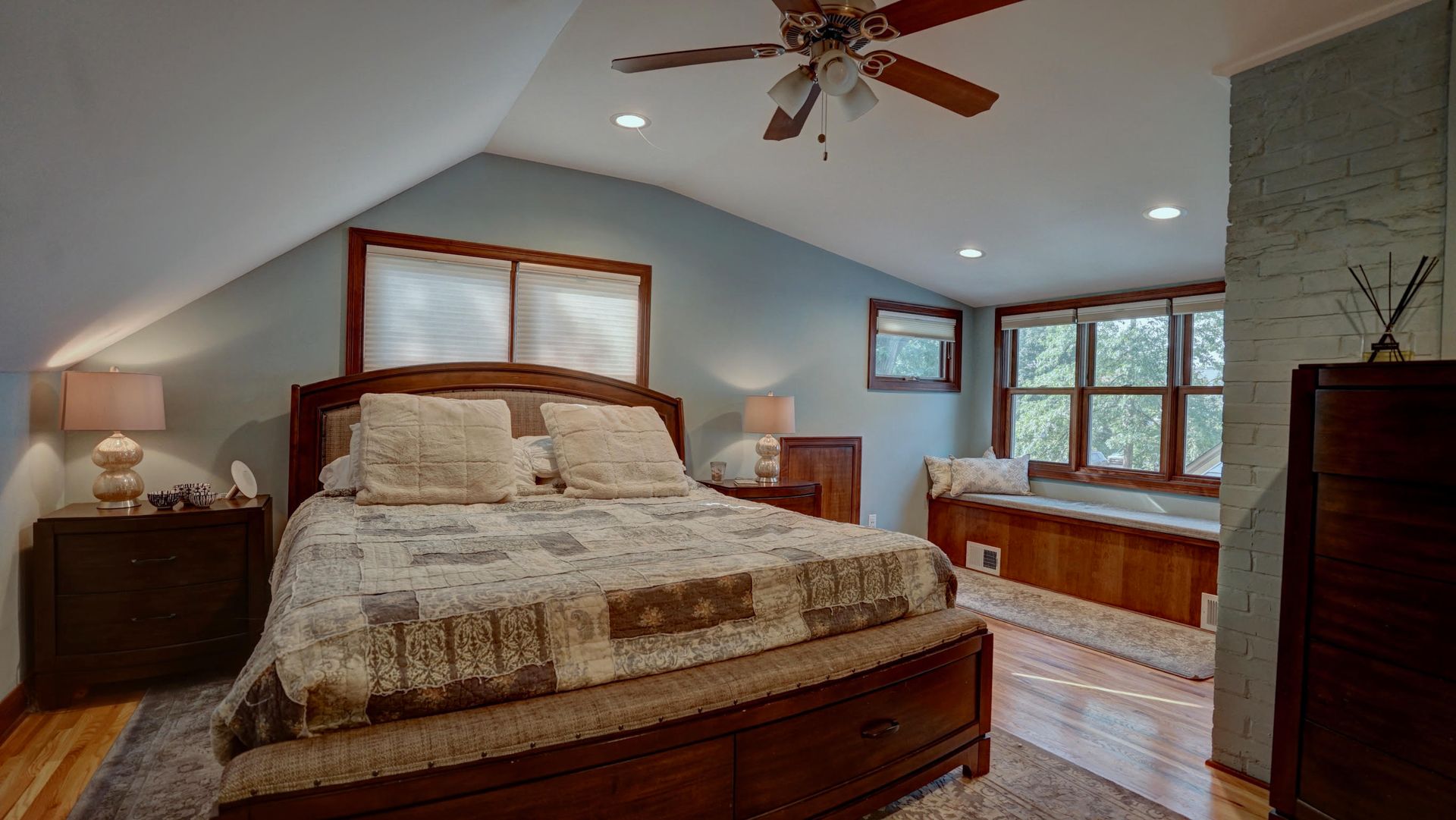 Bedroom with angled ceiling, bed with comforter, window seat, wood furnishings, and light blue walls.