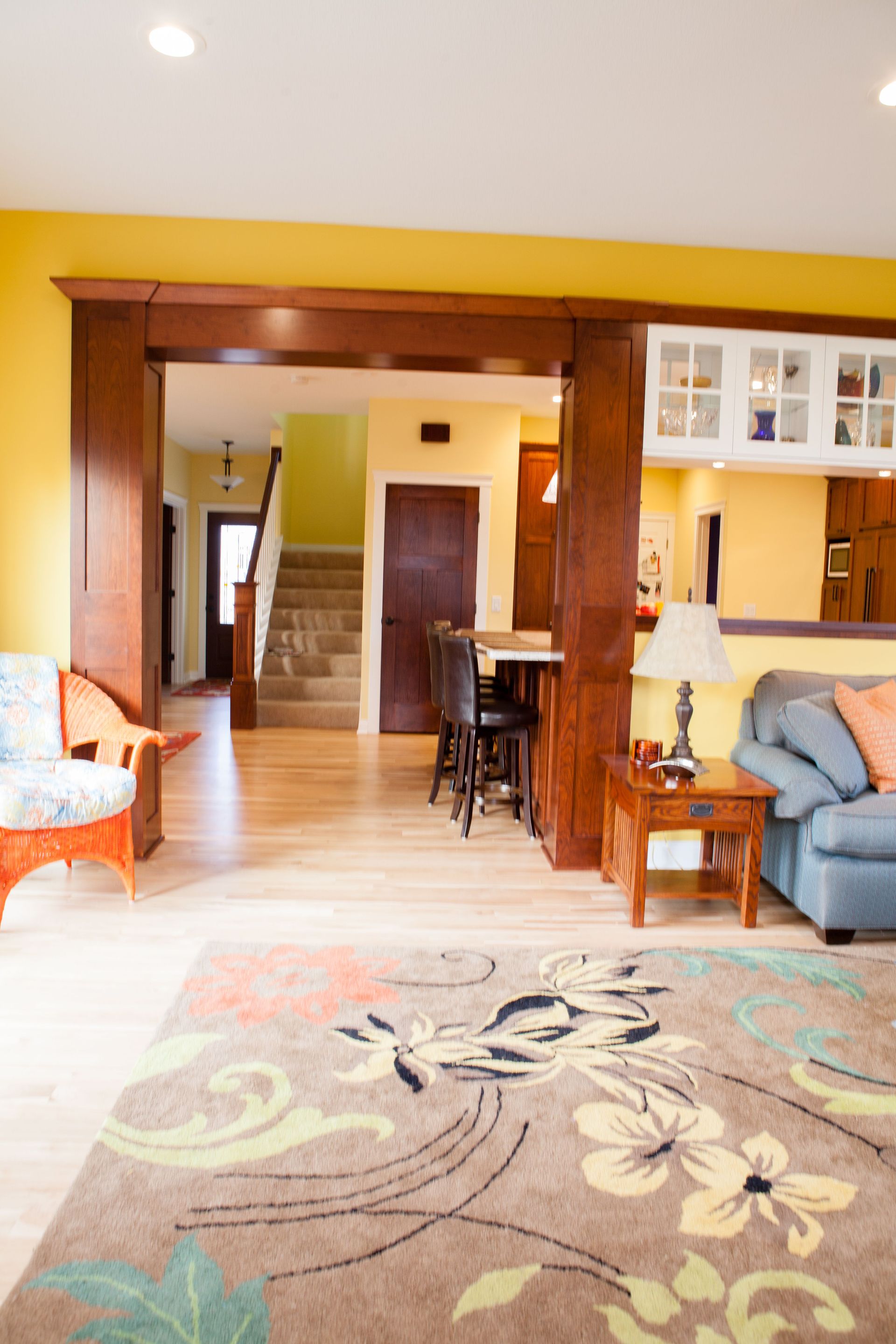 Living room with floral rug, yellow walls, and a view into a kitchen and staircase.