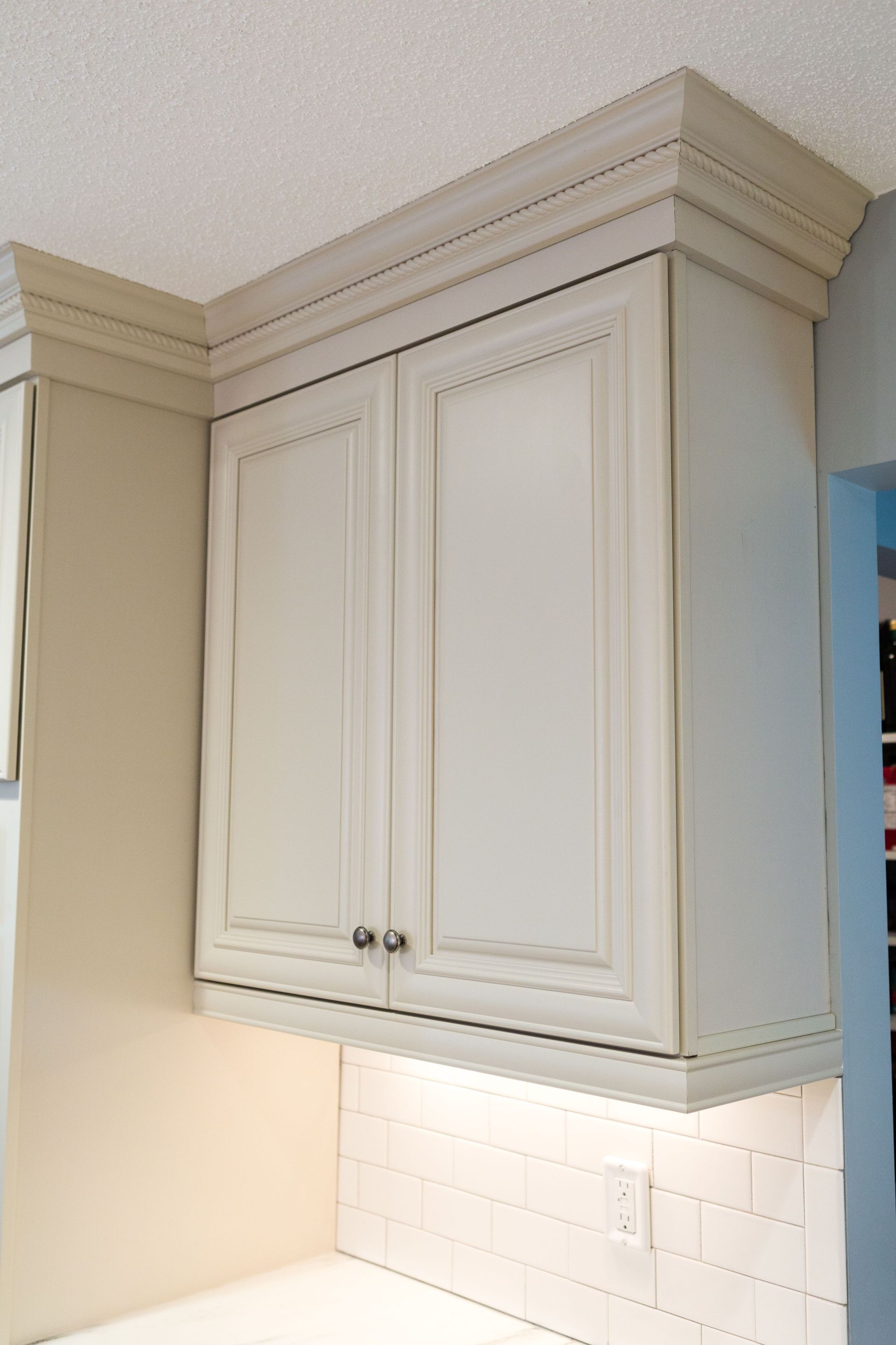 Cream-colored kitchen cabinet with crown molding. White tile backsplash and recessed lighting.