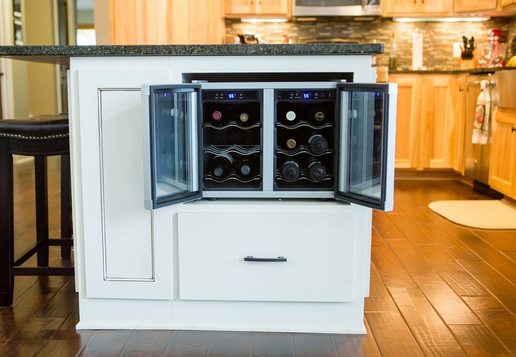 Wine coolers built into a white kitchen island with open glass doors and a dark granite countertop.