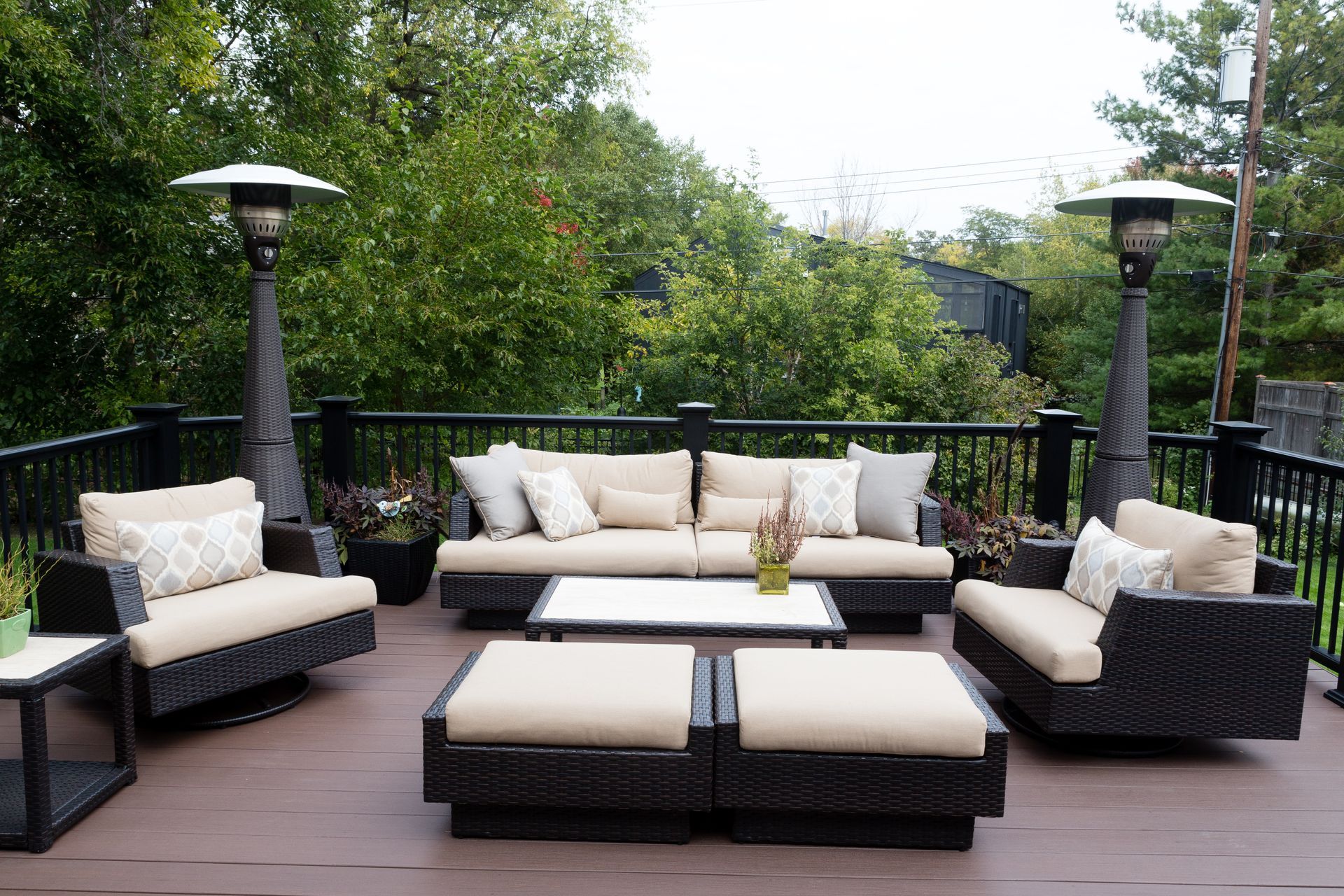 Outdoor patio with beige furniture, two heaters, and a black railing with a green backdrop.