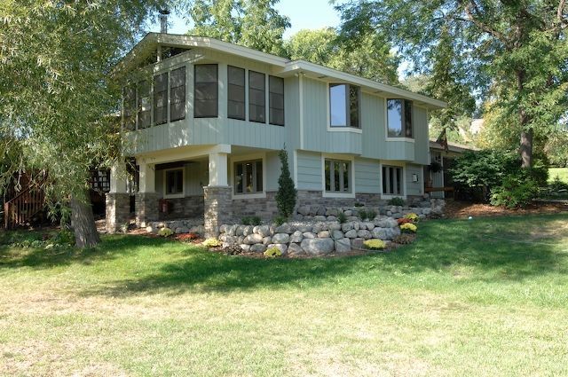Two-story green house with stone base, screened porch, and surrounding trees on a sunny day.