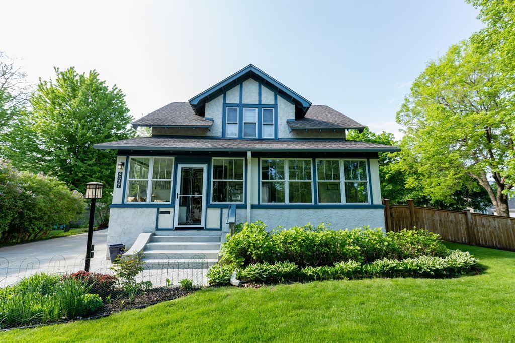 Two-story blue and white house with porch, set on a green lawn with bushes and trees.
