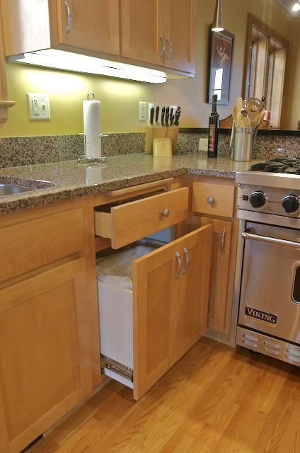 Kitchen with open cabinet, revealing a pull-out trash can. Wooden cabinets and granite countertop.