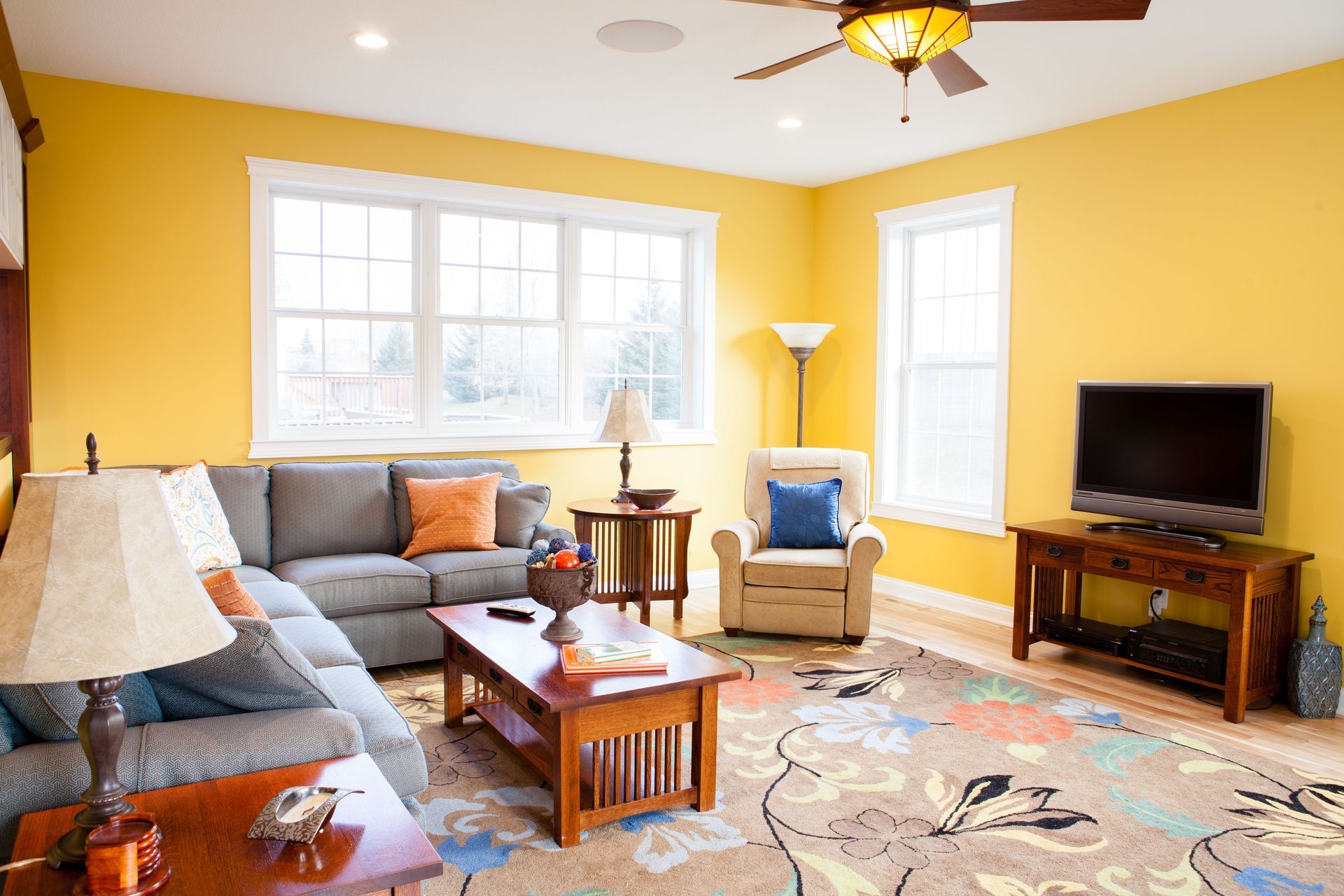 Yellow-walled living room with grey sectional, brown furniture, TV, rug, and ceiling fan.