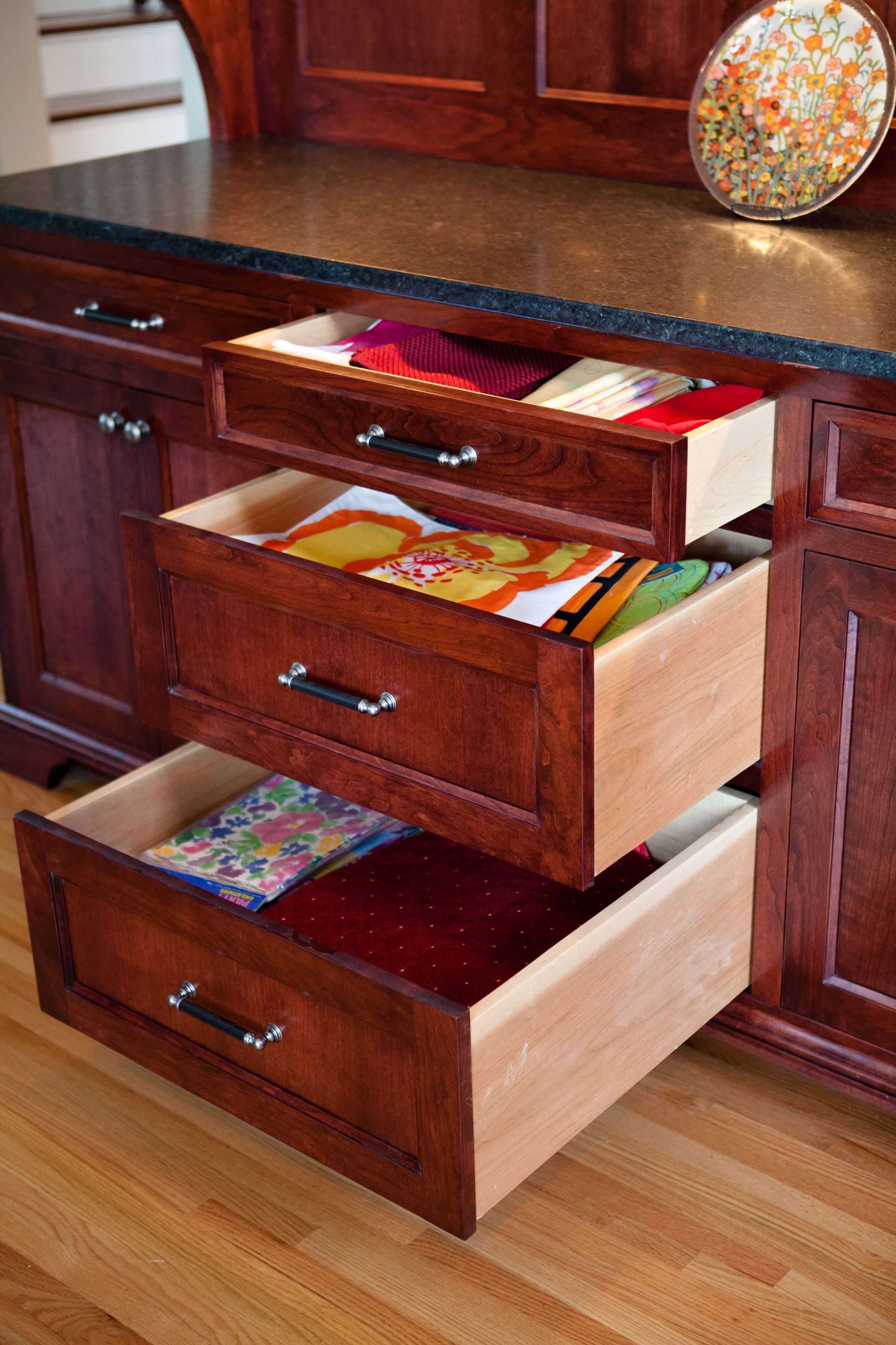 Three open kitchen drawers filled with colorful textiles. Dark wood cabinets, light wood floor.