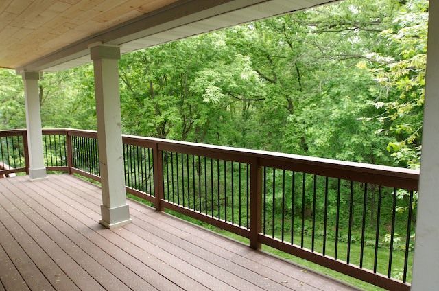 Covered porch with brown decking, railing, and white columns overlooking a green forest.