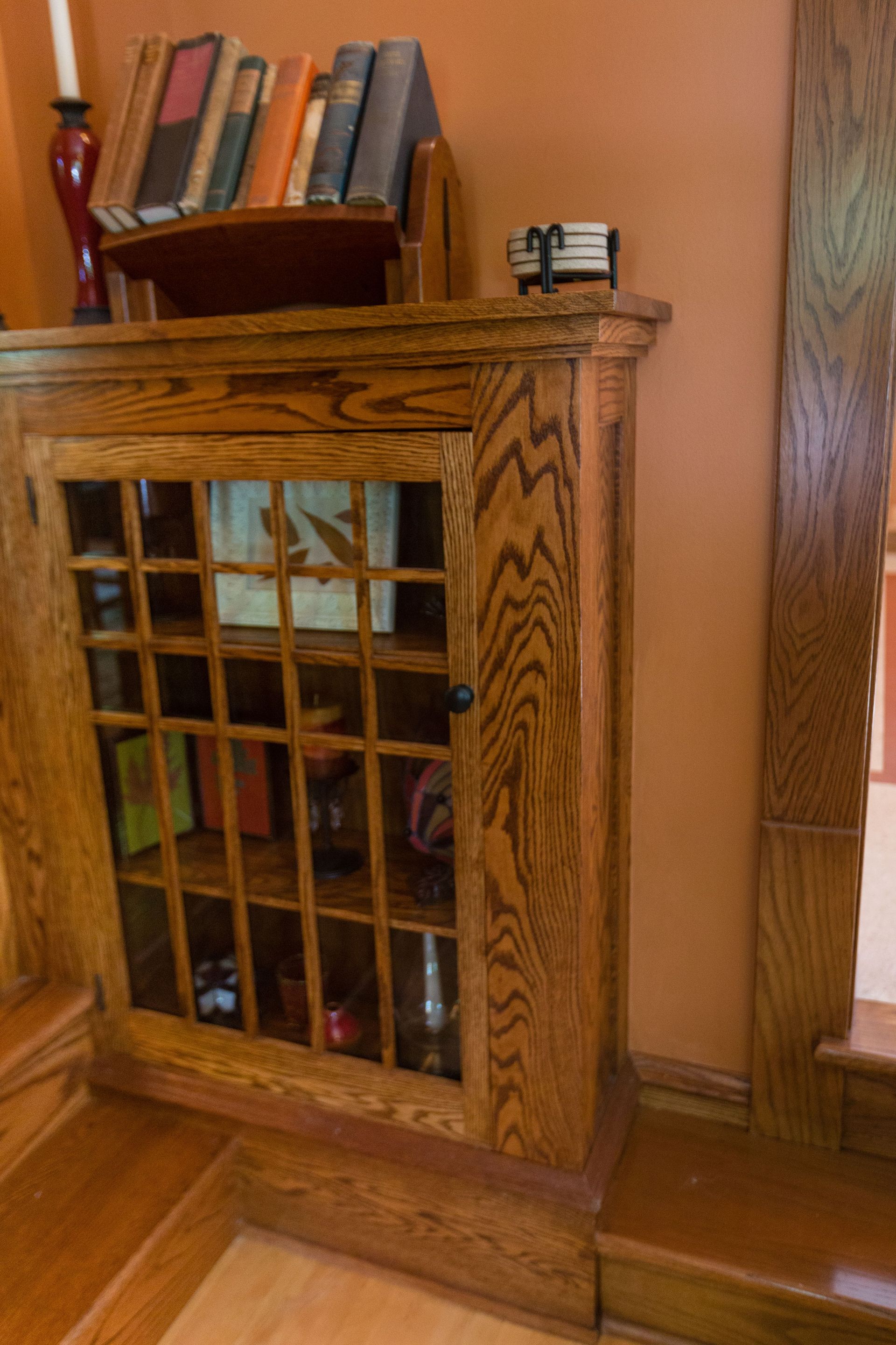 Wooden cabinet with glass grid door, holding books and objects; set in a room with orange walls and wooden trim.