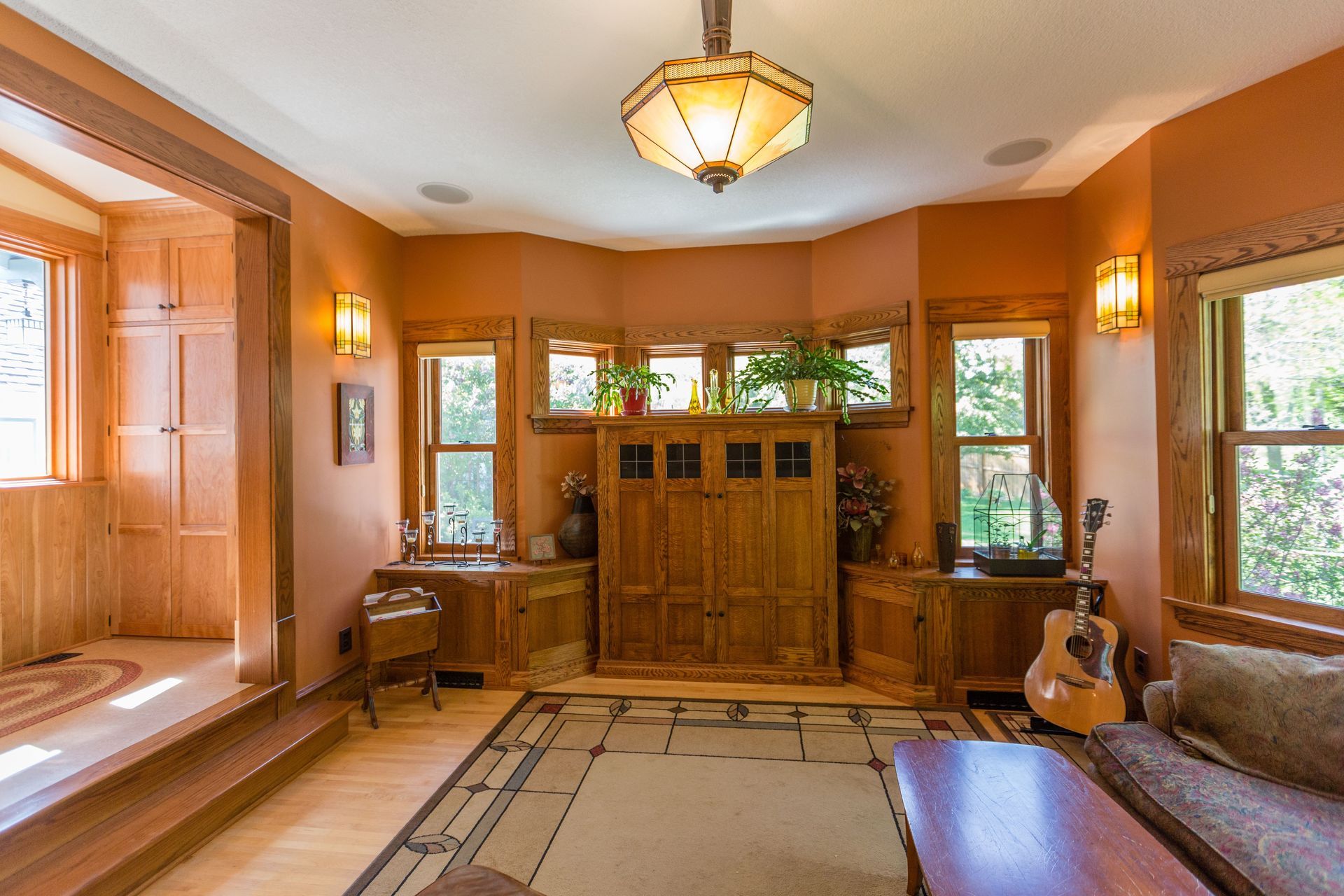 Warm-toned room with wood paneling, windows, and an acoustic guitar. Light-orange walls, and a central wooden cabinet.