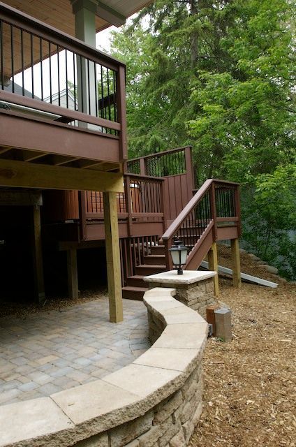 Wooden deck with stairs, brown railing, and a curved stone wall. Backyard setting with trees.