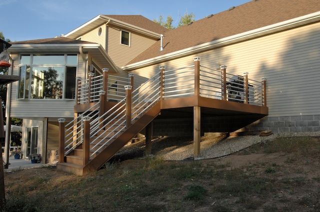 Wooden deck with stainless steel railings attached to a two-story beige house with a glass-walled room.
