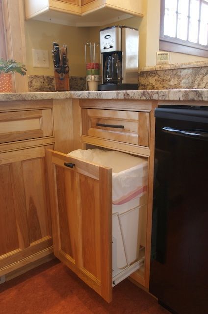 Open cabinet reveals a pull-out trash bin in a kitchen with black appliances and light wood cabinetry.