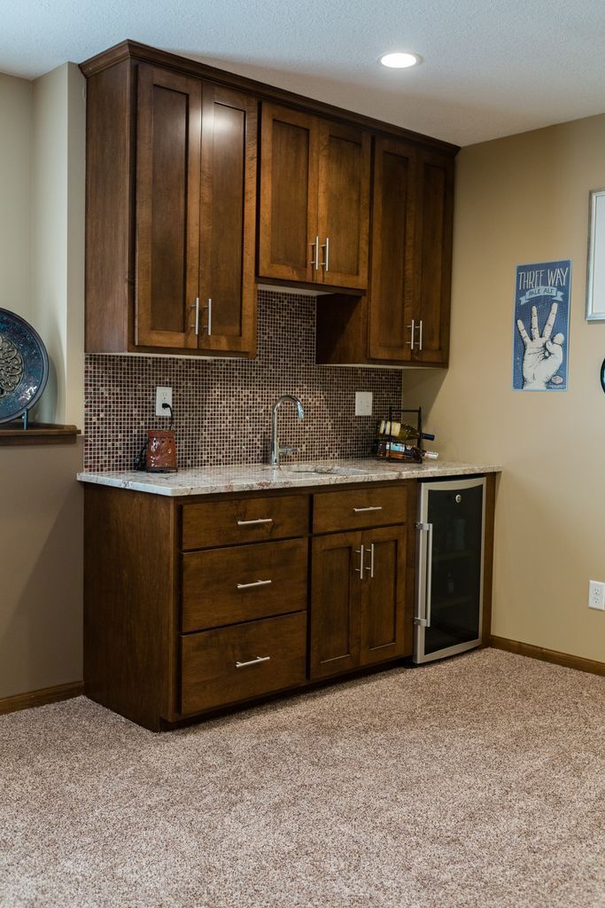 Dark wood built-in bar with cabinets, drawers, and a beverage cooler, against a beige wall and carpet.