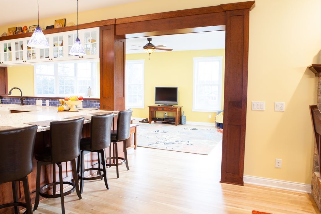 Kitchen with breakfast bar, dark brown stools, opening into a living room with a TV and yellow walls.