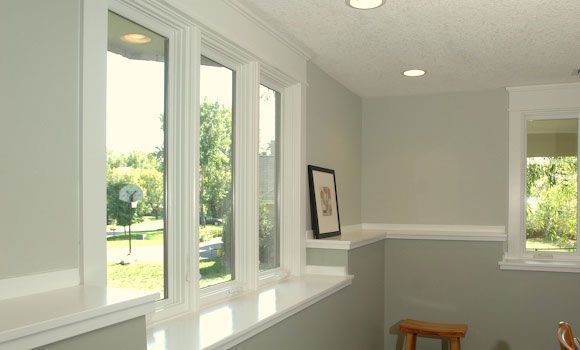 Light-filled room with white-trimmed windows, light gray walls, and a stool; exterior view of green trees.