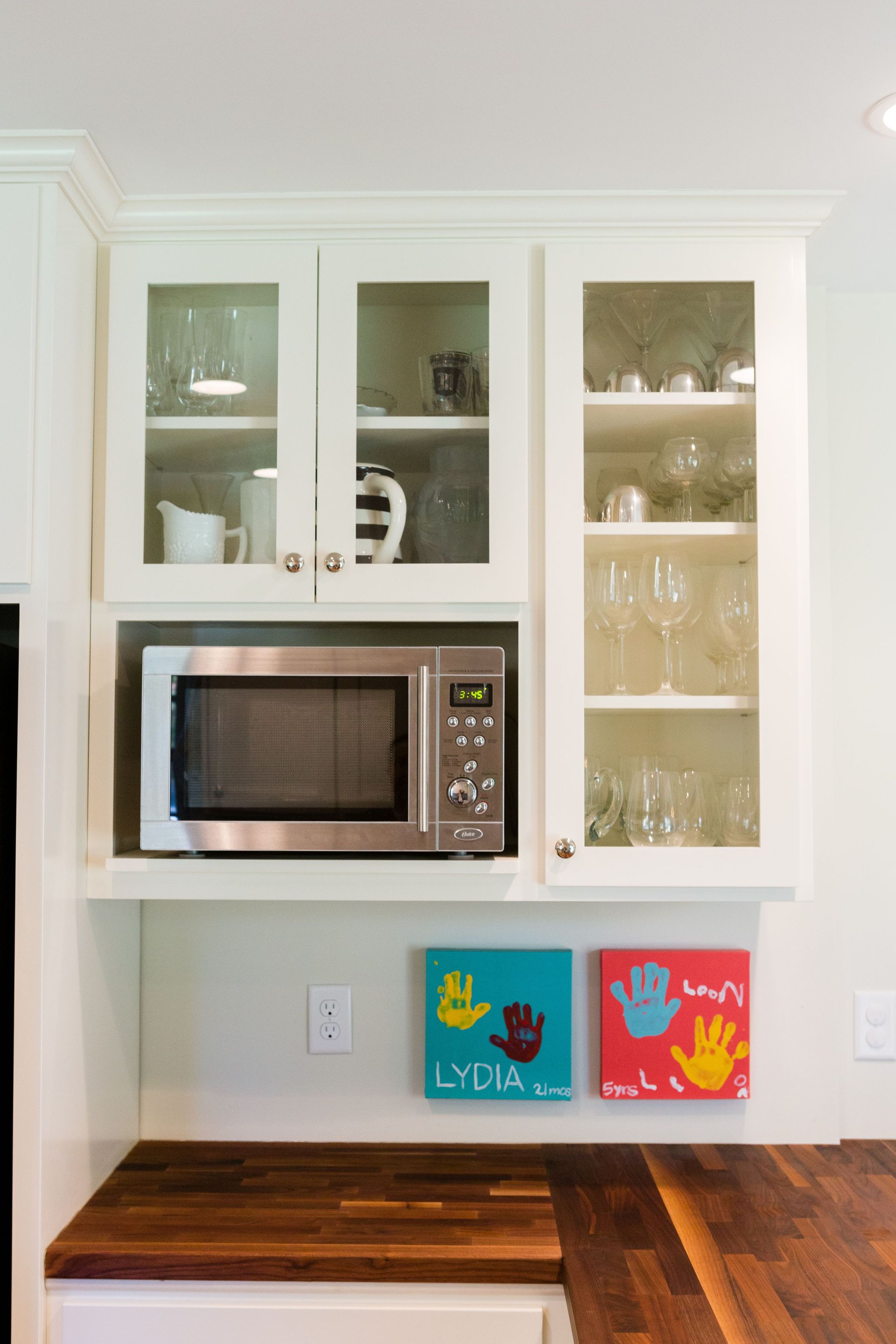 Built-in microwave under a cabinet with glass doors, adorned with children's handprint art, set on a wooden countertop.