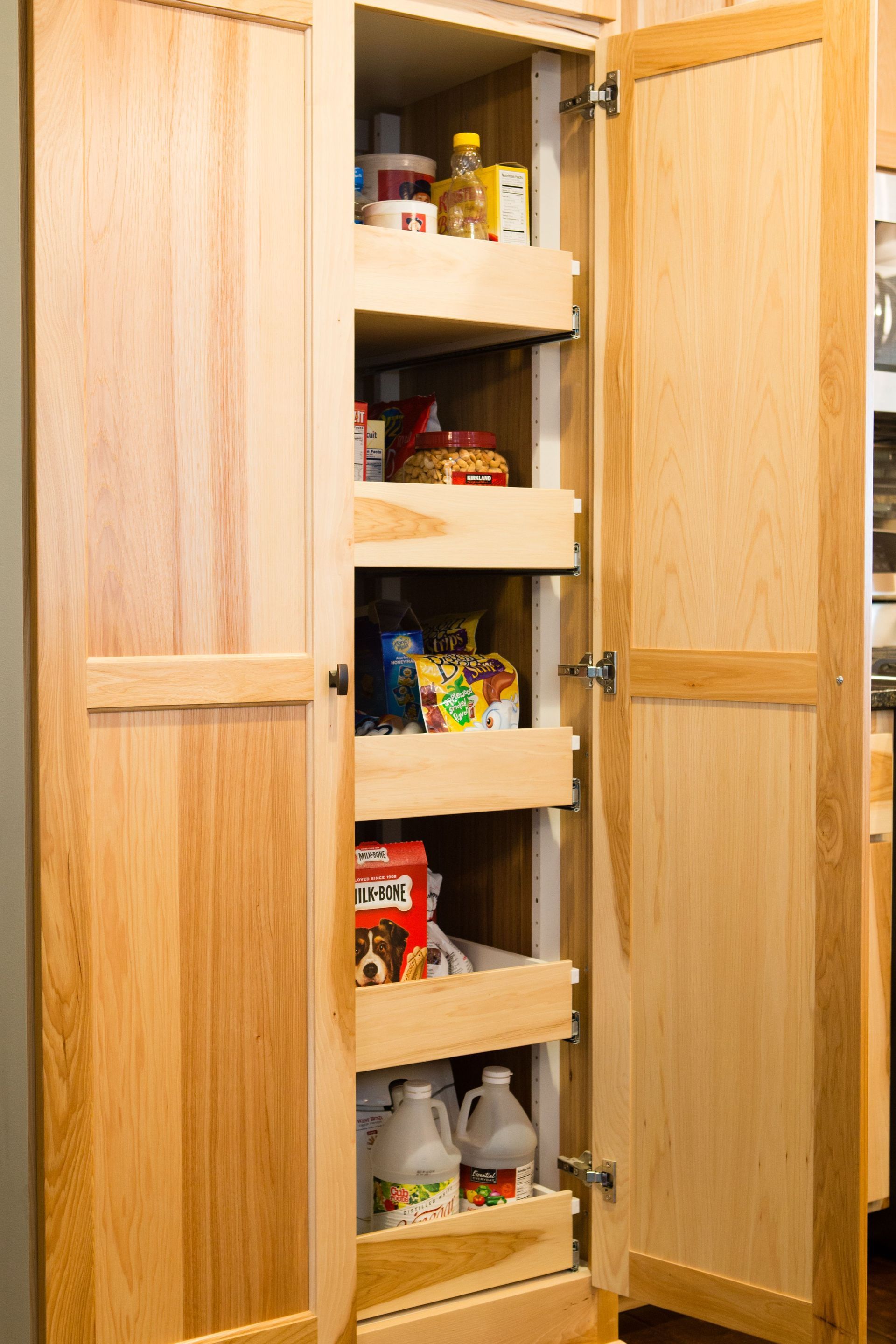 Wooden pantry cabinet with pull-out shelves filled with various food items and cleaning supplies.