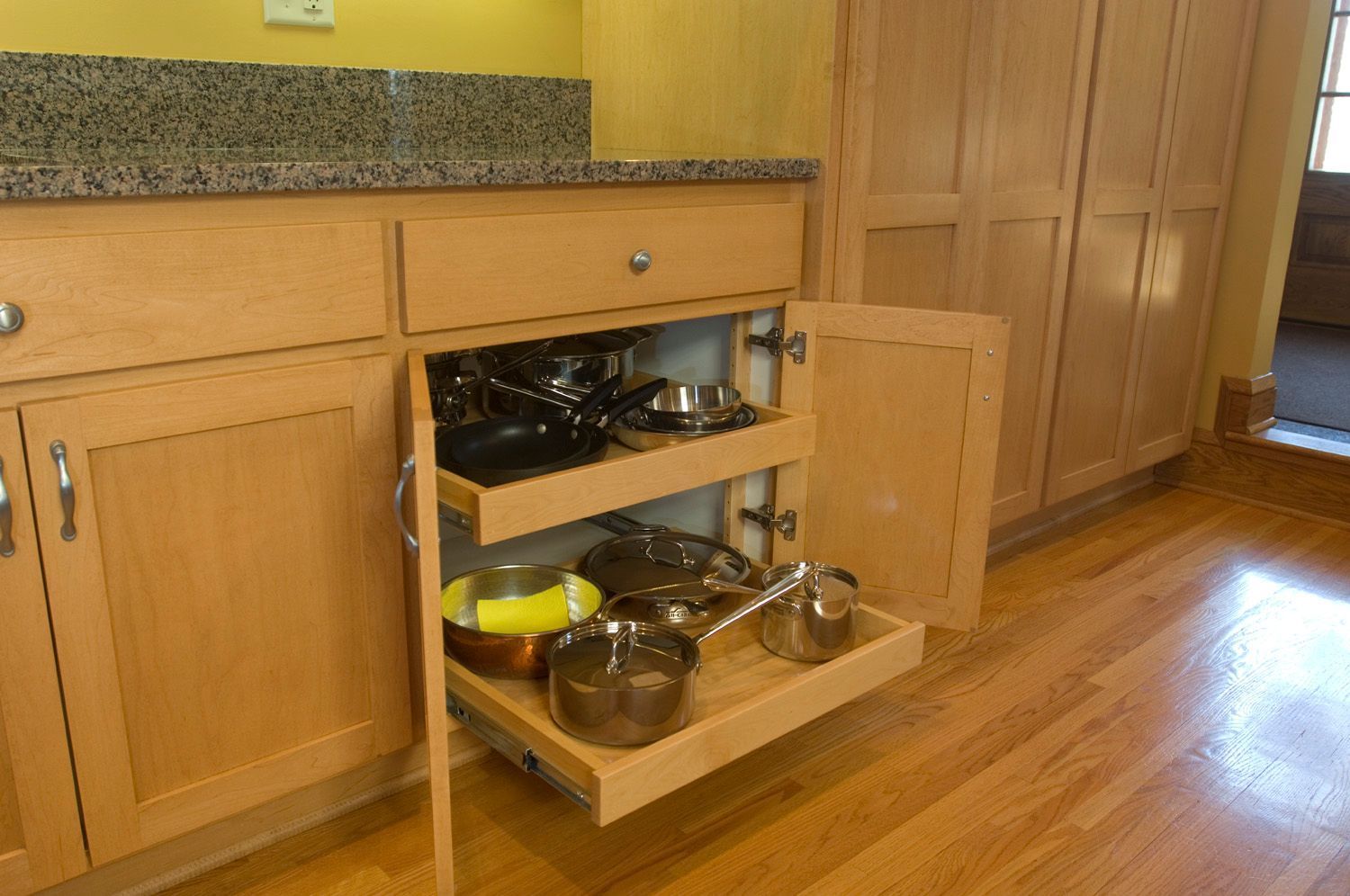 Kitchen cabinet with pull-out shelves holding pots and pans. Light wood cabinets, granite countertop, hardwood floors.