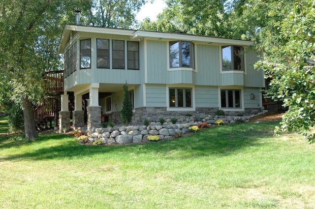 Two-story light blue house with stone base, bay windows, and columns, surrounded by green grass and trees.