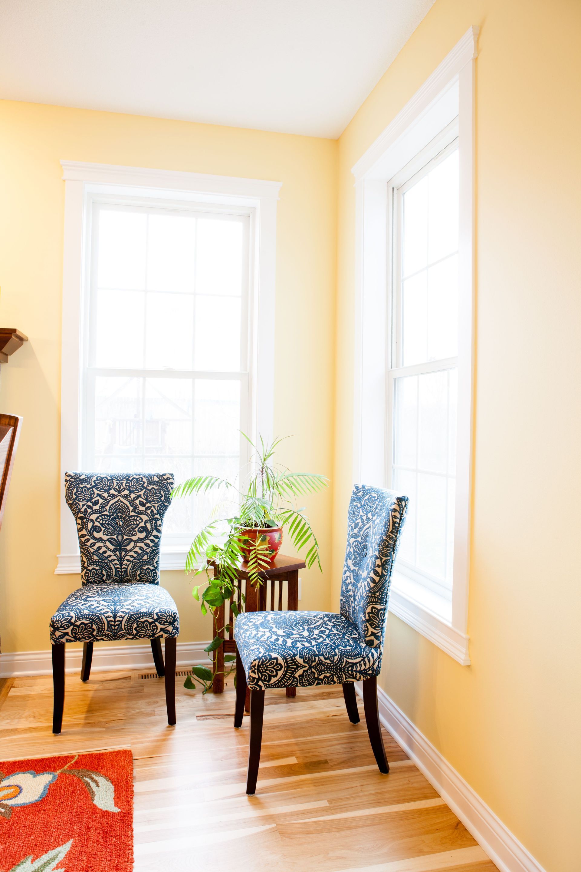 Two patterned chairs near a small table with a plant, placed between two windows in a yellow room.