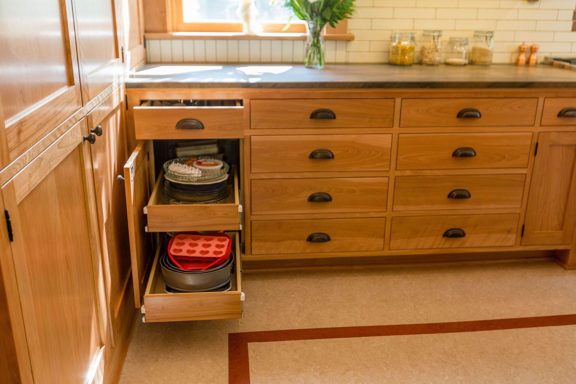 Wooden kitchen cabinets, pull-out drawers holding cake pans, and countertop with jars and window.