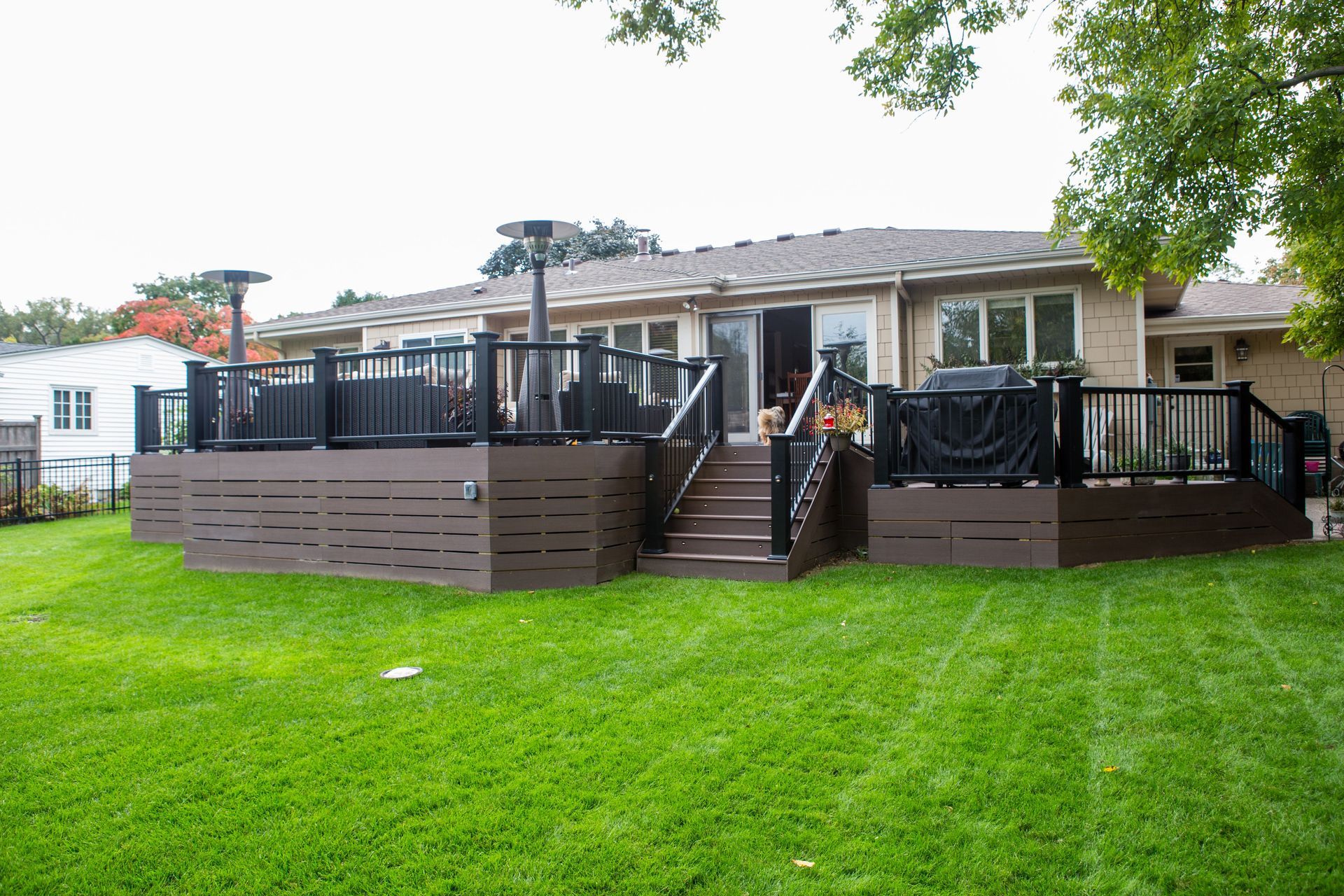 A multi-level wooden deck with black railings and brown siding on a grassy backyard, attached to a beige house.