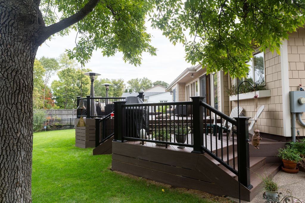 A backyard with a brown deck, black railing, and grill; green grass and trees surround.