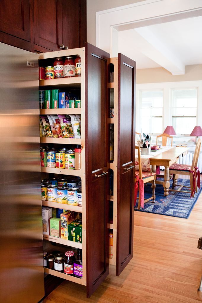 Narrow pull-out pantry cabinet with shelving full of food items, open next to a refrigerator, kitchen setting.