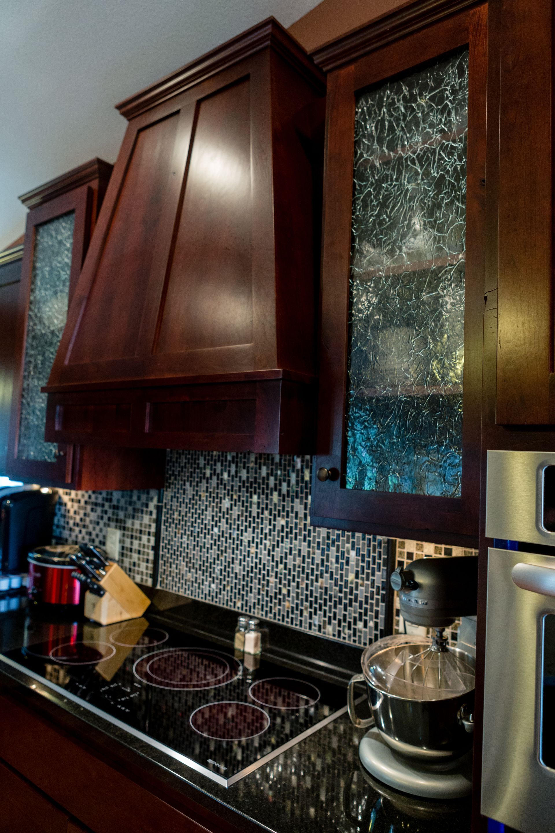 Kitchen with dark wood cabinets, range hood, and mosaic backsplash above a black stovetop.