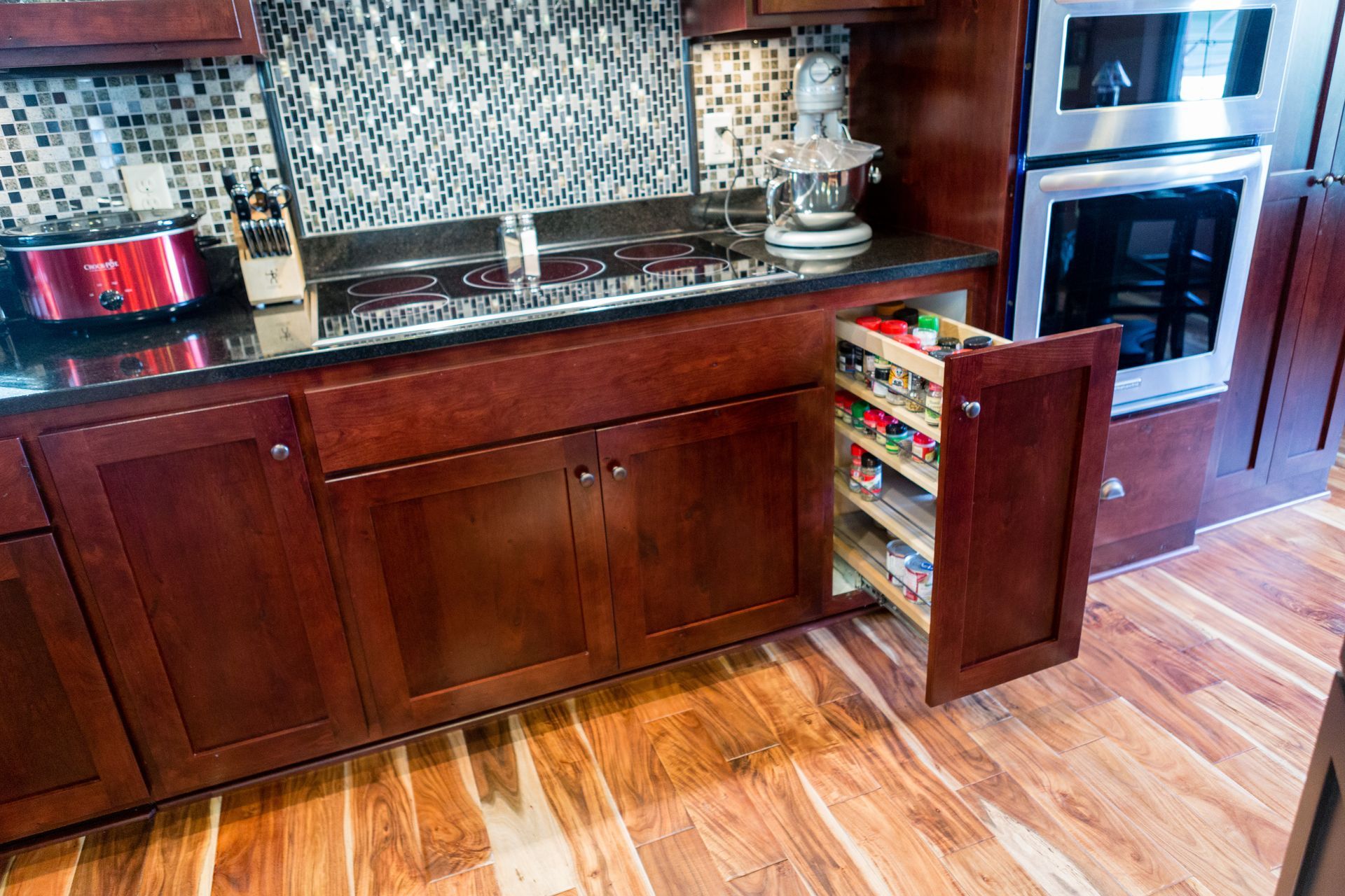 A kitchen with wooden cabinets, black countertop, and pull-out spice rack; oven to the right, wood floor.