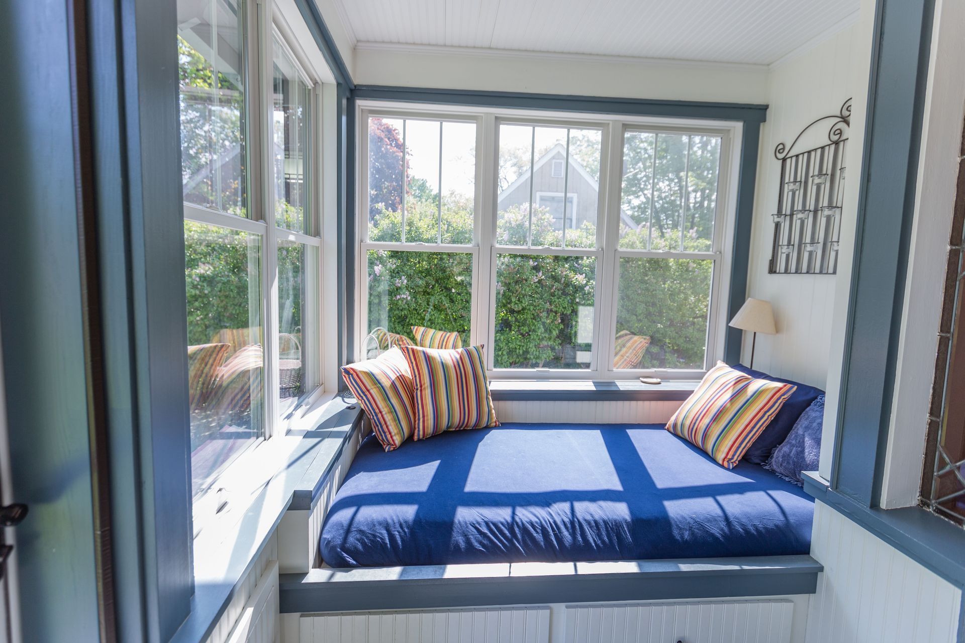 Cozy window seat with blue cushion and striped pillows, surrounded by windows overlooking a lush garden.