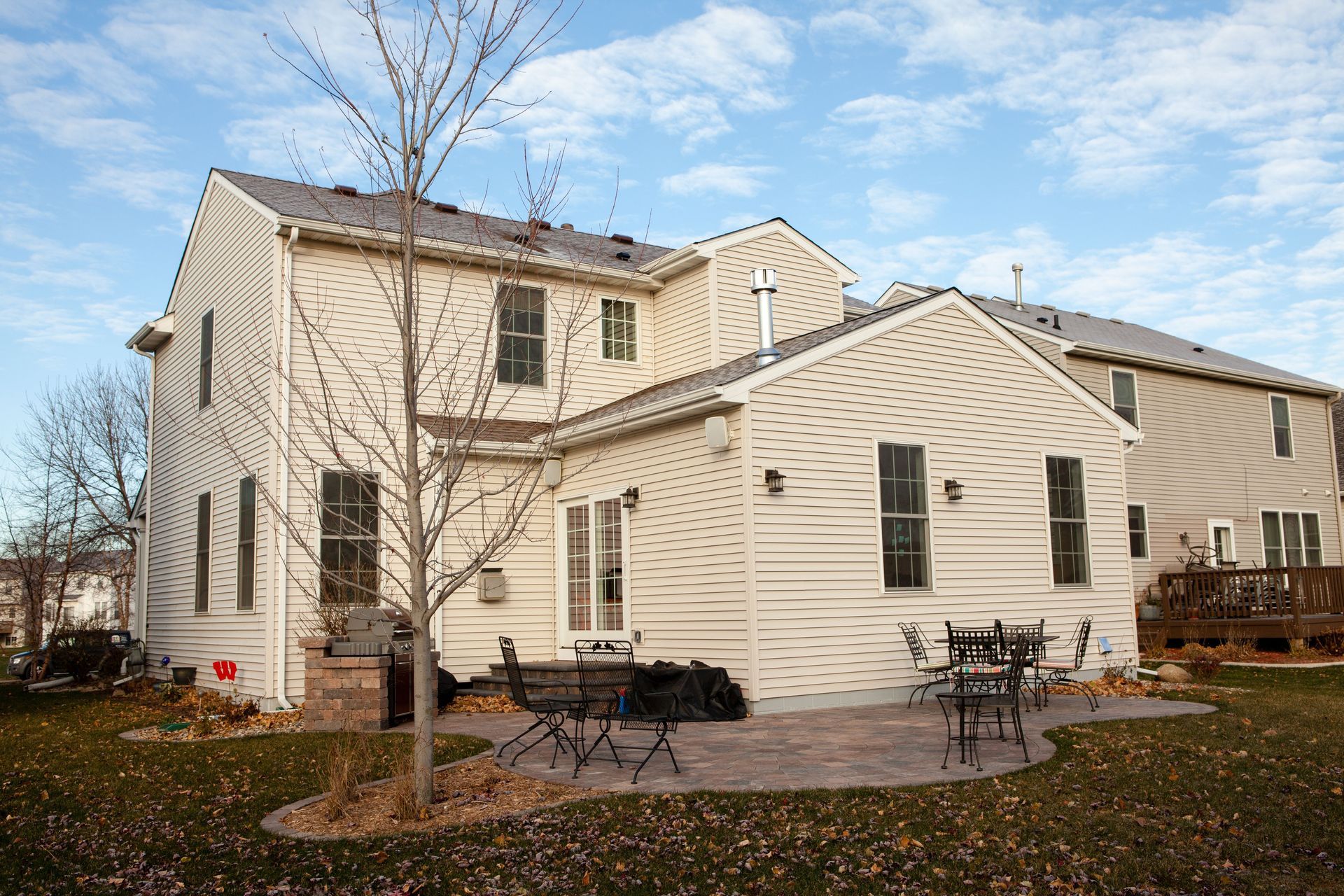 Beige two-story house with a patio, chairs, and bare tree in the yard against a blue sky.