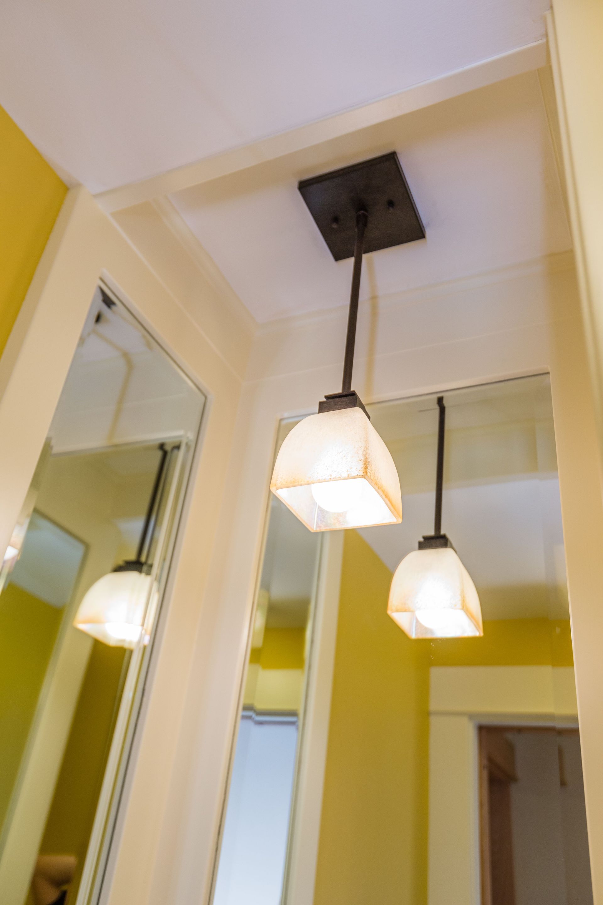 Two pendant lights with white shades hanging from a dark, rectangular fixture. Mirrors and yellow walls are visible.