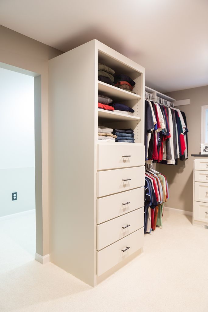Tall white closet organizer with shelves and drawers in a bedroom. Clothes are stored on shelves and hung on rods.