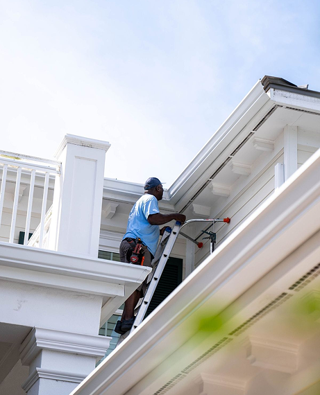 Man on ladder cleaning gutters of a white Little Rock, AR house on a sunny day.
