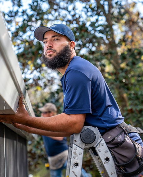 Man on ladder installing gutter, in Little Rock, AR. Another worker in the background. Outdoors setting.
